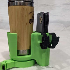 May include: A green 3D-printed cup holder with a bamboo travel mug and two black phone cases. The travel mug has a silver lid and a tractor graphic. The holder has adjustable straps and a swivel base.