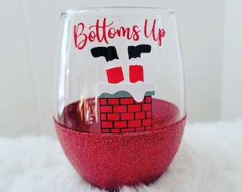 Op de afbeelding: Een helder glazen wijnglas met een rode glitterbodem. Het glas heeft een witte vinyl sticker met een afbeelding van Sinterklaas ondersteboven in een schoorsteen met de tekst "Bottoms Up".