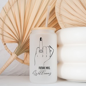 Puede incluir: Un vaso de vidrio esmerilado blanco con un dibujo de línea negra de una mano haciendo el dedo medio con un anillo en el dedo anular. El texto "FUTURE MRS. Williams" está impreso debajo de la mano.