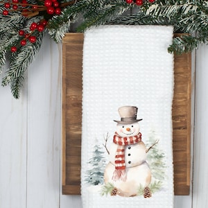 May include: White kitchen towel with a watercolor illustration of a snowman wearing a red and white striped scarf and a black top hat. The snowman is standing in front of a snowy forest scene.
