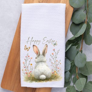Könnte beinhalten: Weißes Küchenhandtuch mit Waffelstruktur und einem Aquarelldesign mit einem weißen Hasen, Blumen und Schmetterlingen. Die Worte "Happy Easter" stehen oben. Das Handtuch liegt auf einem Holzschneidebrett.