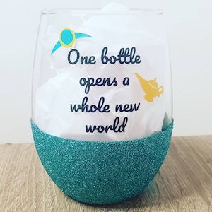 May include: A teal green wine glass with glitter on the bottom half. The glass has a quote that reads "One bottle opens a whole new world" with a gold genie lamp graphic.