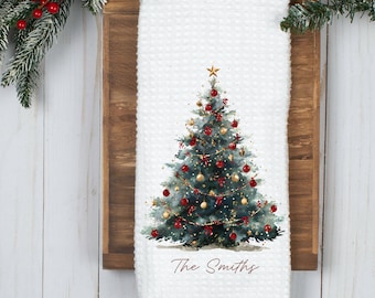 Peut inclure: Un torchon de cuisine blanc à texture gaufrée orné d'une illustration aquarelle d'un sapin de Noël décoré de boules rouges et dorées, d'une étoile dorée et de guirlandes lumineuses, avec le texte The Smiths imprimé en écriture brune.