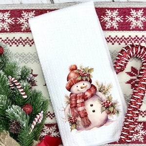 May include: White kitchen towel with a watercolor illustration of a snowman wearing a red hat and scarf. The snowman is surrounded by holly and berries. The towel is on a red and white patterned background.