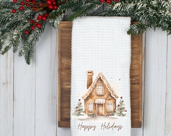 May include: White kitchen towel featuring a gingerbread house illustration with the text "Happy Holidays". The house is brown with white icing and red candy decorations. There are two green trees on either side of the house.