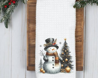 以下が含まれることがあります： 白いワッフル織りのキッチンタオル。水彩画の雪だるまがシルクハットとスカーフを着用し、クリスマスツリーとオーナメントが描かれています。 タオルは木の背景に表示され、常緑樹の枝と赤いベリーが隅にあります。