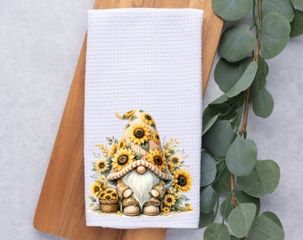 May include: A white kitchen towel featuring a gnome holding a basket of sunflowers. The gnome is wearing a brown hat adorned with sunflowers. The towel has a waffle weave texture.