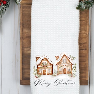 Puede incluir: Toalla de cocina blanca con tejido de gofres con una ilustración de acuarela de dos casas de jengibre con tejados cubiertos de nieve y vegetación festiva. Las palabras "Merry Christmas" están escritas en una elegante escritura. Un artículo de temporada.
