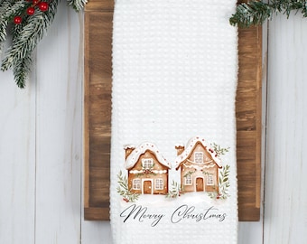 May include: White waffle-weave kitchen towel with a watercolour illustration of two gingerbread houses with snow-covered roofs and festive greenery. The words "Merry Christmas" are written in elegant script below. A seasonal holiday item.