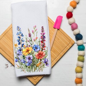 May include: White kitchen towel with a floral design featuring blue, yellow, and red wildflowers. The towel is folded and placed on a wooden chopping board with a pink spatula.