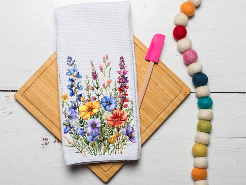 May include: White kitchen towel with a floral design featuring blue, yellow, and red wildflowers. The towel is folded and placed on a wooden chopping board with a pink spatula.