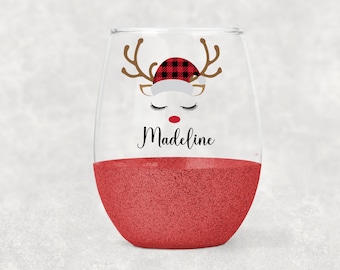 Op de afbeelding: Een rood glitterglas zonder steel met een wit rendiergezicht dat een rood-zwarte geruite kerstmanmuts draagt. Het rendier heeft gesloten ogen en een rode neus. De naam "Madeline" staat onder het rendier gedrukt.