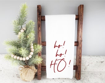 Può includere: Un asciugamano da cucina bianco con testo rosso che dice "ho! ho! HO!" appeso a un portasciugamani in legno. In primo piano c'è un piccolo albero di Natale artificiale con perline bianche.