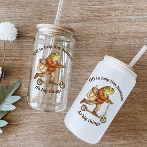 Puede incluir: Dos vasos de cristal con tapas de bambú y pajitas transparentes. Uno es de cristal transparente y el otro de cristal esmerilado. Ambos tienen un gráfico de una rana con chaqueta marrón, llevando una bolsa y montando un patinete con el texto Off to help tiny humans do big things.