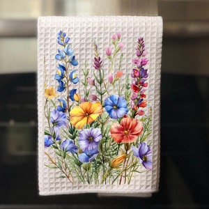 May include: A white waffle weave kitchen towel with a colourful floral design. The flowers are painted in shades of blue, yellow, orange, pink, and purple. The towel is hanging on a kitchen cabinet.