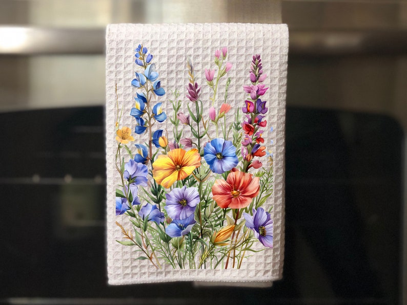 May include: A white waffle weave kitchen towel with a colourful floral design. The flowers are painted in shades of blue, yellow, orange, pink, and purple. The towel is hanging on a kitchen cabinet.