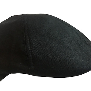 May include: A black flat cap made of a textured fabric. The cap has a slightly curved brim and a rounded crown.