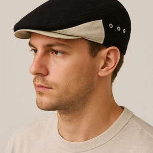 May include: A black flat cap featuring a beige brim and a beige accent panel on the side. The side of the cap includes three small silver-toned metal eyelets arranged in a horizontal row, providing ventilation and a distinct design detail.