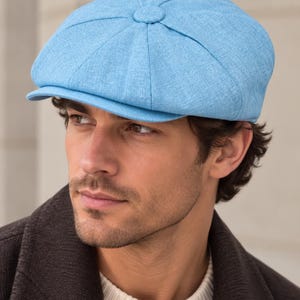 May include: A light blue newsboy cap featuring a structured eight-panel design with a central fabric-covered button on the crown and a short, stiff brim. The cap is crafted from a textured fabric with a visible weave pattern throughout the material.