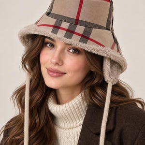 May include: A beige bucket hat featuring a red, black, and white plaid pattern. The hat is lined with a soft, light beige faux fur material and includes matching ear flaps with long, dangling beige knit ties hanging down on both sides.
