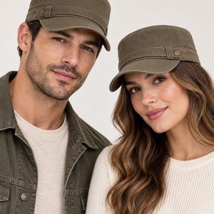 May include: Two olive green military-style cadet caps featuring a flat top, a short curved brim, and a decorative side strap secured with a small metallic button. The caps are constructed from a solid-colored fabric with visible stitching details.