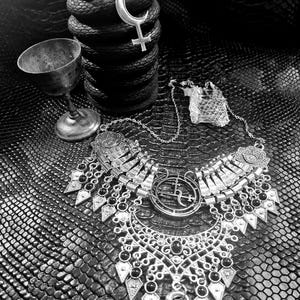 May include: A monochrome image showing a lit candle on a textured, coiled candle. A silver chalice and a detailed necklace with a snake pendant are visible. The necklace features black detailing and geometric designs.
