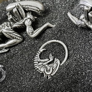 May include: Close-up of silver-colored Alien-themed jewelry. The image features a hinged nose ring with an Alien face design, and several Alien figures. The jewelry is on a textured, dark surface. A logo is visible.