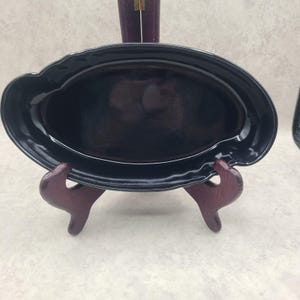 May include: A glossy black oval ceramic serving platter with a scalloped edge, displayed on a dark wooden stand. The dish features a smooth, reflective surface and a deep basin, providing a clean and simple aesthetic for serving various food items.