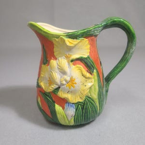 May include: A ceramic pitcher featuring a vibrant orange background with a raised, three-dimensional yellow iris flower design. The handle is shaped like a green stem, and the base is decorated with green leaves and a small blue flower bud.