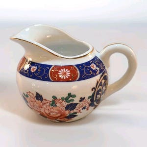 May include: Cream-colored ceramic creamer with a round body and a handle. The creamer features a blue band with floral and scroll designs, and a red and white floral medallion. The rim is trimmed with gold.