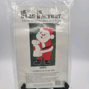 May include: A complete flag kit featuring a Santa Claus design. The kit includes a blank flag, instructions, and extra fabric for design. The packaging reads "The Flag Factory" and "Create Your Own Beautiful Flag!"