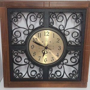 Peut inclure: Horloge murale carrée avec un cadre en bois brun foncé et un motif en fer forgé noir. Le cadran de l'horloge est doré avec des chiffres et des aiguilles noirs. C'est un élément de décoration intérieure.