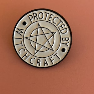 May include: A circular enamel pin with a white background and black border. The centre features a black pentagram inside a circle. The text "PROTECTED BY WITCHCRAFT" is embossed in black capital letters around the perimeter, separated by two black dots.