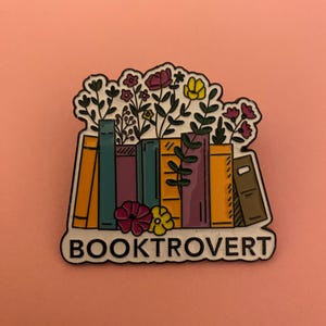 May include: An enamel pin with a black outline, depicting a stack of books with flowers. The pin has the word "BOOKTROVERT" in white text on a white background. The background is a solid peach color.