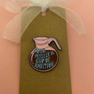 May include: An enamel pin in the shape of a coffee pot, with the text "Pour Myself A Cup of Ambition" in blue. It's attached to a brown tag with a white ribbon. The background is a pale pink.