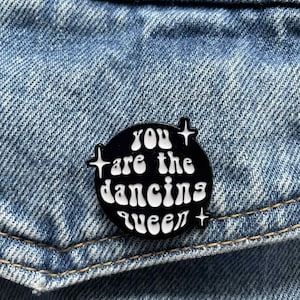 May include: A black enamel pin with the text "You are the dancing queen" in white, set against a denim background. The pin is round with star-shaped accents. A fun accessory for jackets, bags, or hats.