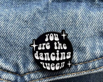 May include: A black enamel pin with the text "YOU are the dancing queen" in white, set against a denim background. The pin has a circular shape with star-like accents.