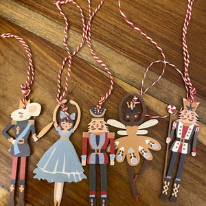May include: Five Christmas tree decorations depicting characters from The Nutcracker ballet. The ornaments include a mouse, a ballerina, a nutcracker soldier, a fairy, and a second nutcracker soldier. Each is made of paper and has a red and white string for hanging.