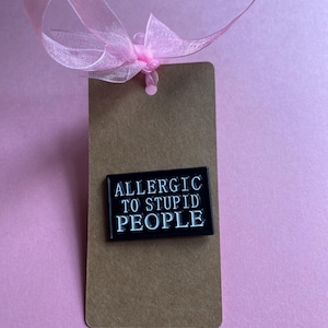 May include: A brown rectangular tag with a black pin that reads "ALLERGIC TO STUPID PEOPLE" in white lettering. A sheer pink ribbon is tied at the top of the tag, set against a pink background.