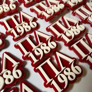 May include: Red and white ΠΛ 1986 pins. The pins feature the Greek letters Pi and Lambda, along with the year 1986 in a bold, stylized font. The pins have a red background with white lettering.
