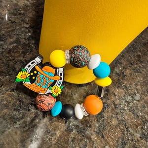 May include: A colorful beaded bracelet with a charm of a cowboy boot and horseshoe. The beads are yellow, orange, brown, black, white, and blue. The charm is brown, yellow, and orange.