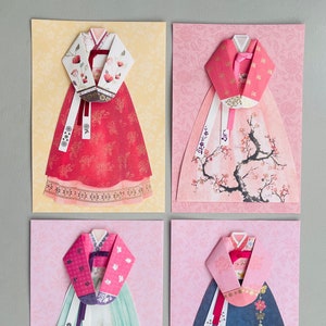 May include: Four cards with origami paper cutouts of traditional Korean hanbok dresses. Each card features a different color and pattern, including red, pink, green, and blue. The dresses are decorated with floral and butterfly motifs.