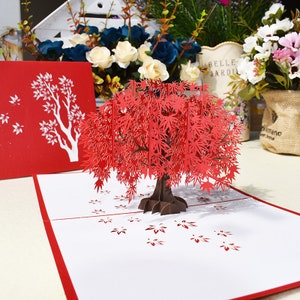 May include: A red 3D pop-up card featuring a detailed papercraft maple tree with leaves falling to the ground. The card is closed and has a red cover with a white silhouette of a tree.