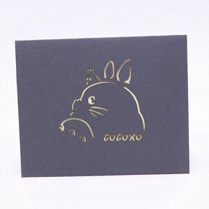 Pu&ograve; includere: Una carta grigia con un ritaglio bianco di Totoro, un personaggio del film Il mio vicino Totoro, con una piccola creatura sulla testa. La parola "Totoro" &egrave; scritta sotto il personaggio.