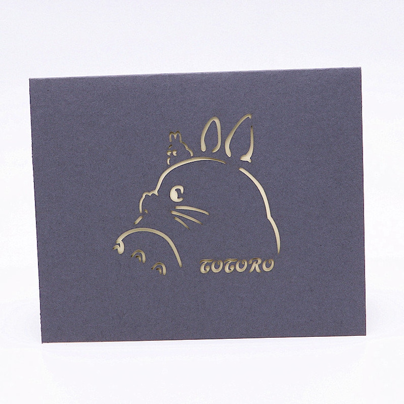 Pu&ograve; includere: Una carta grigia con un ritaglio bianco di Totoro, un personaggio del film Il mio vicino Totoro, con una piccola creatura sulla testa. La parola "Totoro" &egrave; scritta sotto il personaggio.
