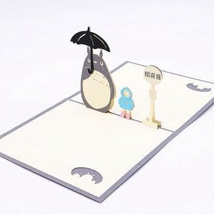 Pu&ograve; includere: Un biglietto pop-up 3D con un personaggio di Totoro grigio e bianco che tiene un ombrello nero, una piccola figura vestita di blu e un cartello che dice "Fermata dell'autobus".