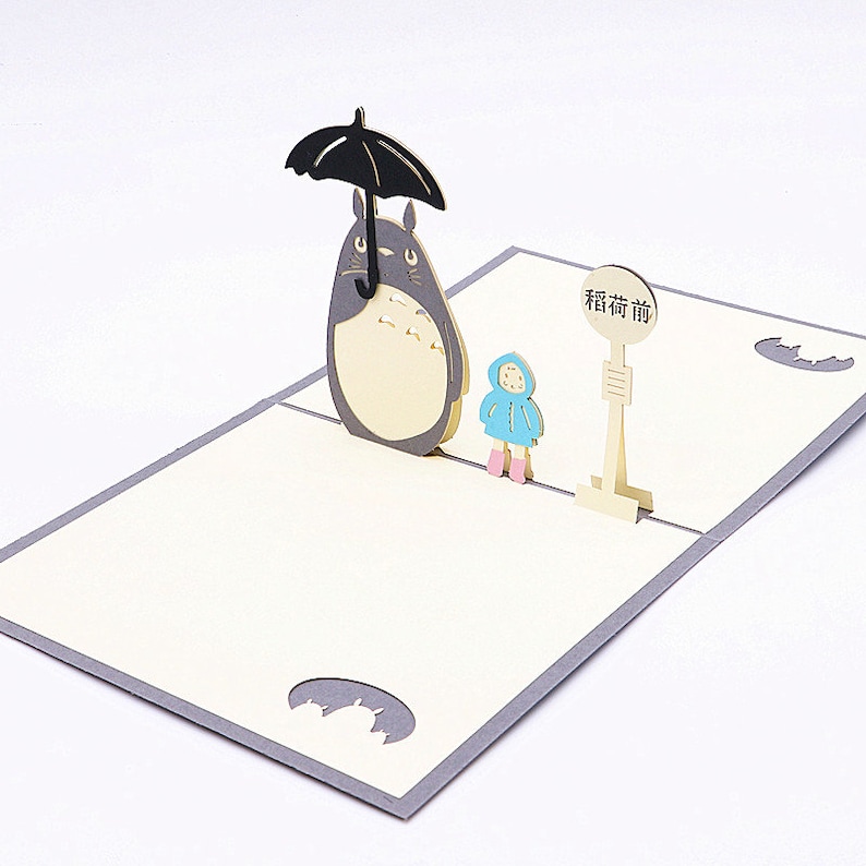 Pu&ograve; includere: Un biglietto pop-up 3D con un personaggio di Totoro grigio e bianco che tiene un ombrello nero, una piccola figura vestita di blu e un cartello che dice "Fermata dell'autobus".