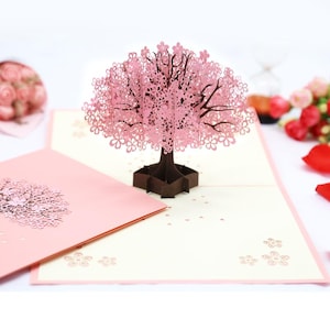 May include: A pink 3D pop-up card featuring a cherry blossom tree with intricate paper cut details. The card is open and the tree is standing upright. The card is on a white background.