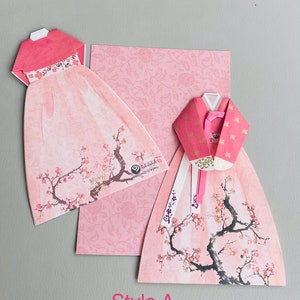 May include: Two pink origami paper Hanbok dresses with a cherry blossom design. The dresses are folded into a three-dimensional shape and have a white ribbon tied around the waist. The background is a pink floral pattern.