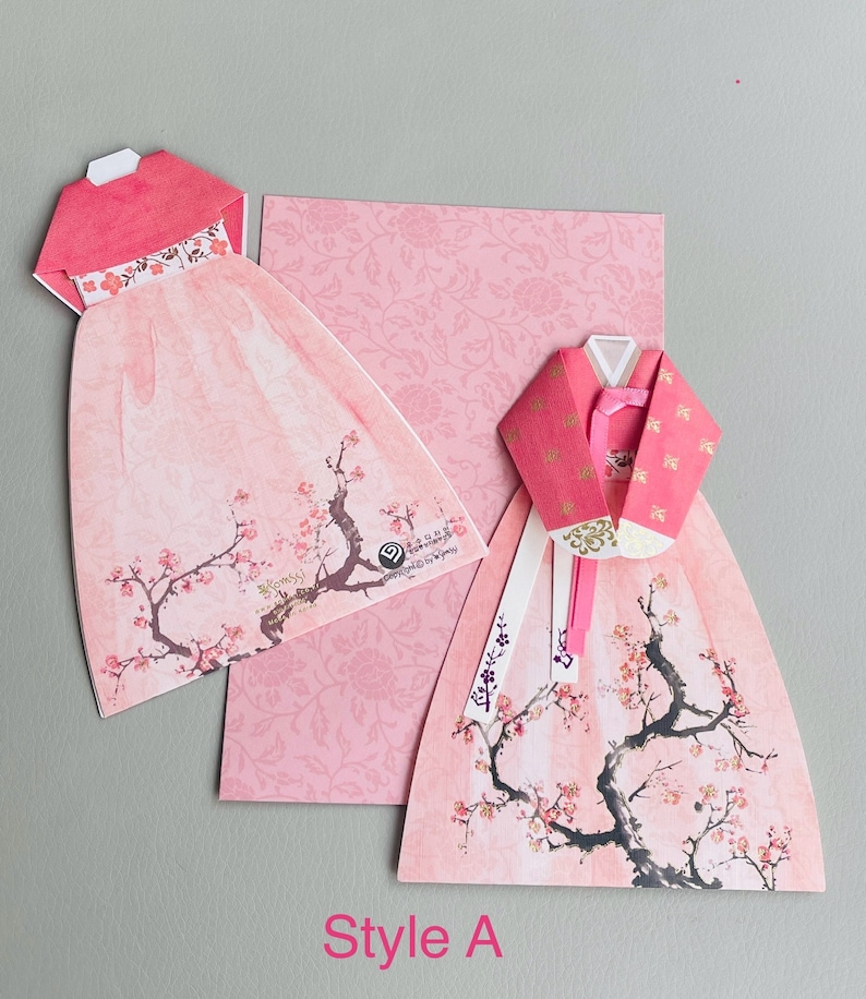 May include: Two pink origami paper Hanbok dresses with a cherry blossom design. The dresses are folded into a three-dimensional shape and have a white ribbon tied around the waist. The background is a pink floral pattern.